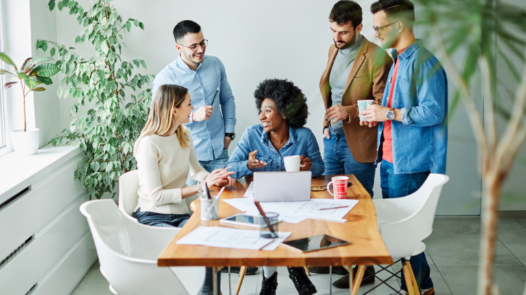 A group of five people, including TA Leaders, are gathered around a wooden table with documents and a laptop, engaged in a discussion. They are in a bright room with plants and natural light.