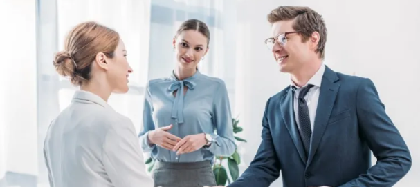 Three colleagues are having a conversation in an office. A woman and a man are shaking hands, sealing what feels like a promising deal, while another woman stands in the background, observing with a smile that brings a sense of warmth to their professional home.