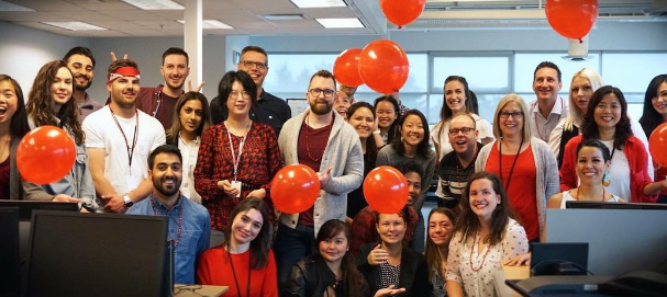 A group of people in an office setting, some holding red balloons, smile for a photo. Computer monitors are visible in the foreground, adding a touch of home to their collaborative space.