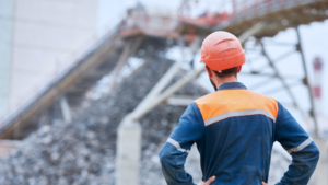 A worker in a hard hat and high-visibility vest stands with hands on hips, measuring success as they look at a conveyor belt system at a construction or industrial site.