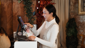 A woman in an apron smiles while using a touchscreen device on a counter, seamlessly handling high volume hiring in a cozy, plant-filled environment.