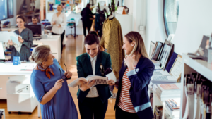 Three women in a stylish office discuss a magazine, while enterprise teams work at desks in the background. The setting, featuring large windows and modern decor, reflects the dynamic environment essential for high volume recruitment.