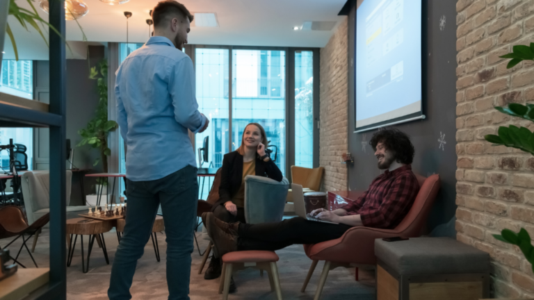 Three people are having a casual conversation in a modern office lounge. One person is standing, and the other two are seated, with a presentation screen visible in the background displaying "Streamlining Hourly Hiring.