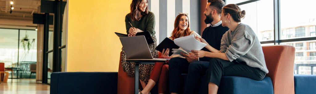 Four colleagues sit on a sofa in an office lounge, engaging in a discussion about job descriptions while holding laptops and documents, with large windows in the background.