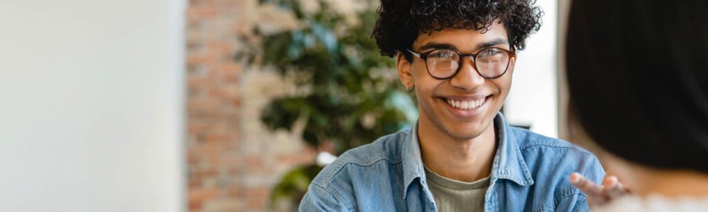 A person with curly hair and glasses smiles while sitting indoors, with another person partially visible in the foreground, embodying the essence of hiring best practices.