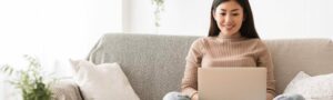 A woman sits on a beige sofa working on a laptop in a bright room, likely sourcing top talent for her company hiring push.