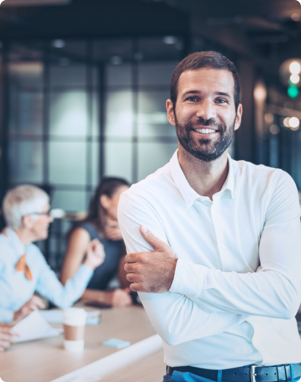 A man with a beard in a white shirt stands smiling with arms crossed in the foreground of an office setting focused on talent acquisition. Three HR leaders are conversing in the background.