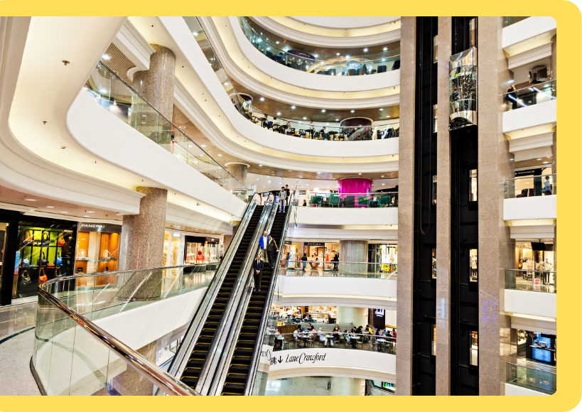 A multi-level shopping mall with glass railings, escalators, and various retail stores. People are visible on different floors and on the escalator, exploring a variety of services.